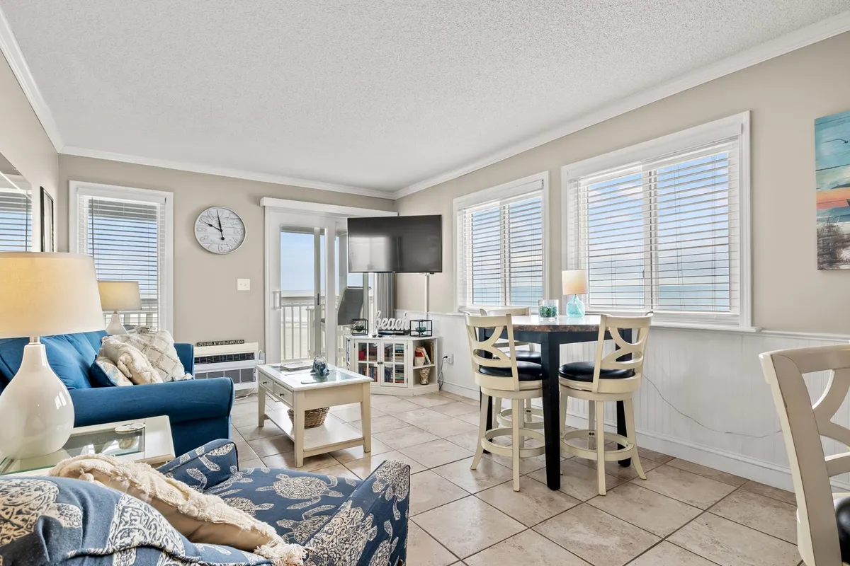 The image showcases a bright, welcoming living space with a plush blue sofa, a spacious dining table perfect for gatherings, and expansive windows framing stunning sunrise ocean views. The room is styled for comfort and togetherness, highlighted by cheerful decor and a wall clock that hints at carefree vacation time.