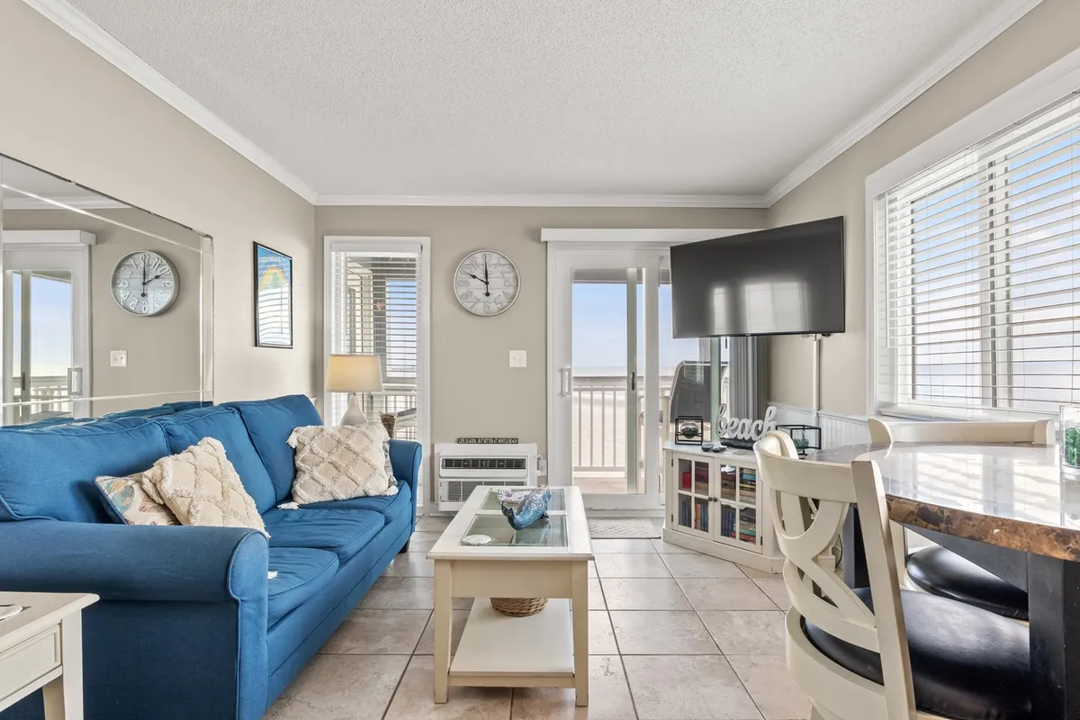 This image captures the open-plan living and dining space, featuring a plush blue couch, a dining table with chairs, large windows, and a TV. Sunlight floods the room through the expansive windows and sliding doors, creating a bright and airy social hub perfect for relaxing, entertaining, or watching a sunset after a day at the beach.