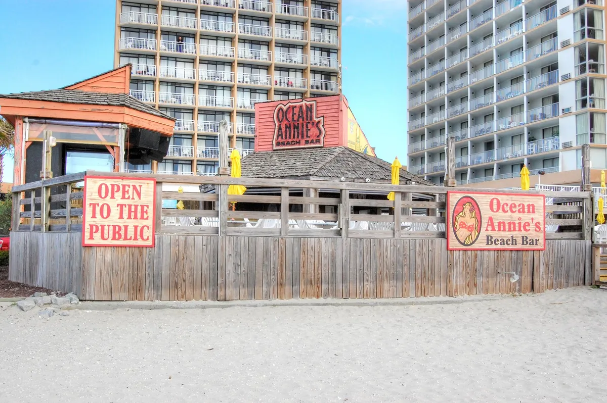 This image showcases Ocean Annie's Beach Bar, featuring the recognizable sign, an inviting wooden deck with railings, and vibrant yellow umbrellas. People are visible socializing and relaxing in a lively, beachfront setting.