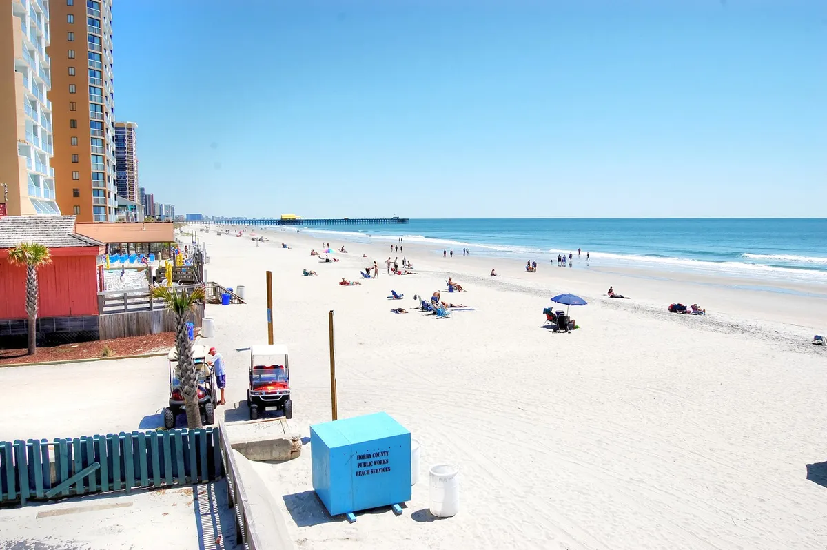 A lively beach scene featuring families lounging on the sand, children building sandcastles, and the ocean stretching out to a nearby pier. The backdrop includes beachfront buildings, reinforcing the proximity of accommodations to the shore. The overall atmosphere is vibrant and family-oriented, perfectly illustrating easy beach access and playful moments.