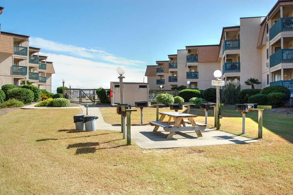 The image features outdoor grill stations, a picnic table, lush greenery, and condo balconies, creating a welcoming space perfect for group barbecues and social gatherings.