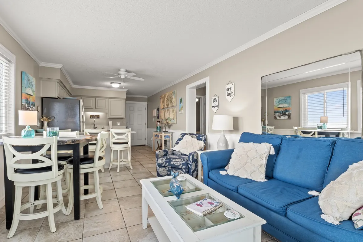 A slightly different angle of the living and dining area, this image highlights a cozy blue sofa, dining table with chairs, vibrant wall art, and a mirror that reflects natural light. The setting feels intimate and inviting, ideal for family gatherings, casual meals, or game nights.