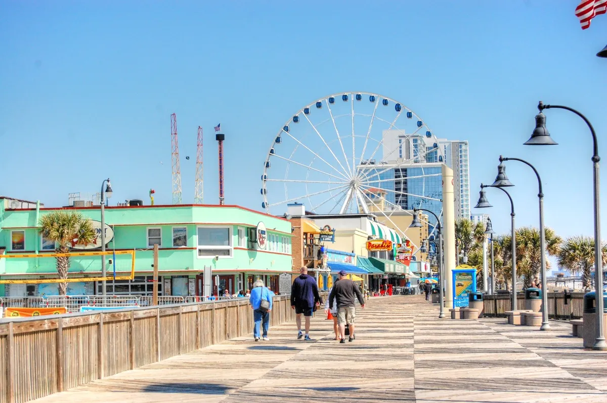 A bustling boardwalk scene with a Ferris wheel towering above colorful buildings, and families strolling along the walk. The lively environment suggests nearby attractions, entertainment, and local experiences that complement beach days with unforgettable outings.