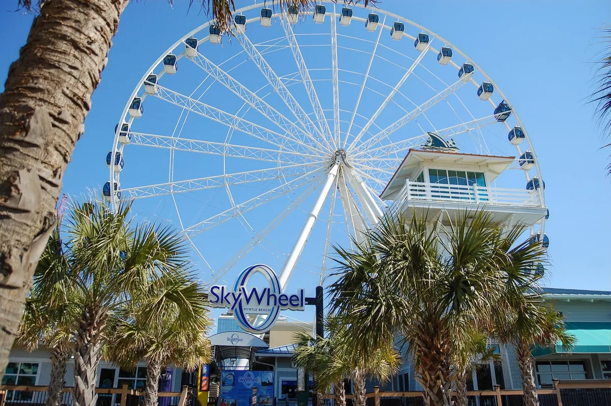 A bright exterior shot of the SkyWheel Ferris wheel, surrounded by palm trees and a ticket booth, capturing the excitement and energy of one of Myrtle Beach's most iconic attractions.