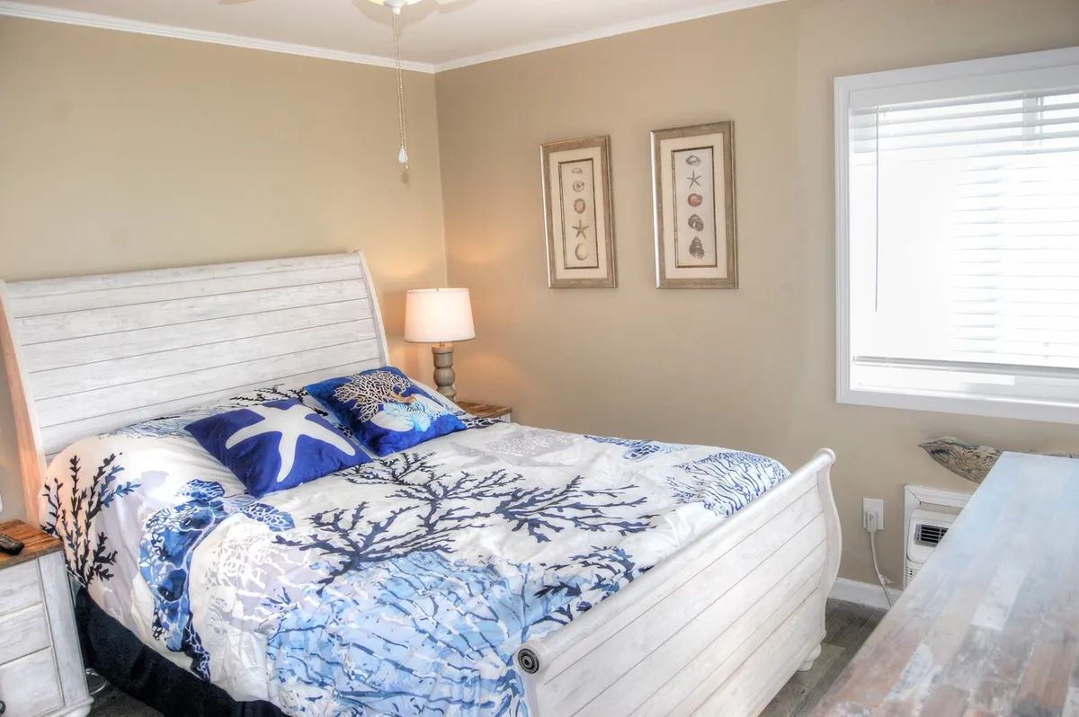 This image features a queen-sized bed dressed in plush coral and sea-themed bedding, accented by a decorative starfish pillow and framed coastal artwork on the wall. A warm bedside lamp casts a tranquil glow, reinforcing the room’s peaceful, beach-inspired décor.