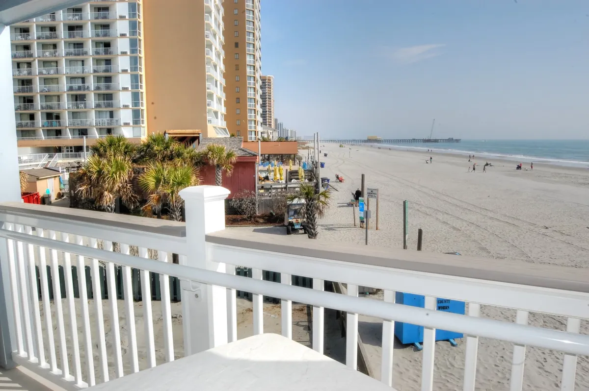 A sweeping balcony view overlooking Shore Drive’s sandy beach, the ocean stretching into the distance, a palm-lined pier, and gentle waves. The image captures the tranquility and romance of watching the sunset or sunrise from a private vantage while connecting directly to the beachside location emphasized in the section.