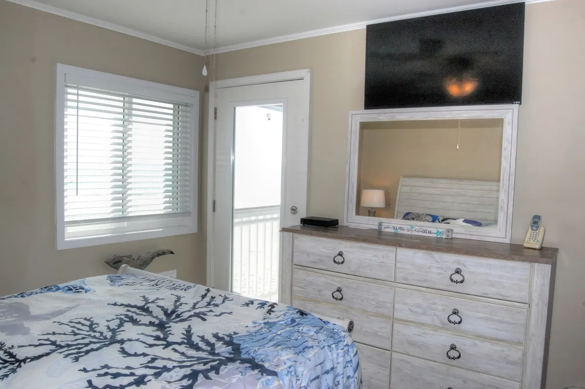 A soothing bedroom scene with a coral-patterned queen bed, a dresser with a mirror, and sunlight streaming through the window. The door to the outside hints at a private, restful escape.
