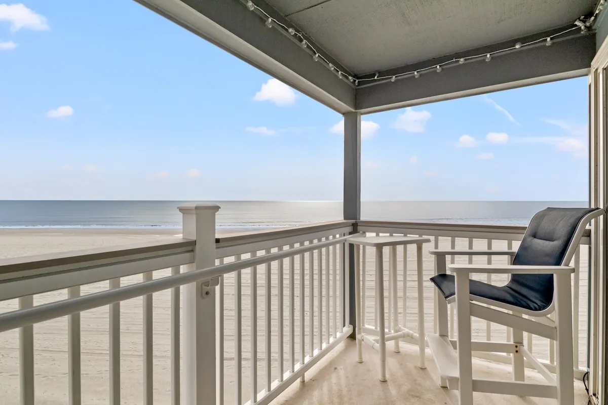 A serene balcony scene featuring a single chair and small table overlooking the beach and ocean. Soft natural light bathes the space, and the view stretches out to the waves, capturing a tranquil and private moment ideal for relaxation.