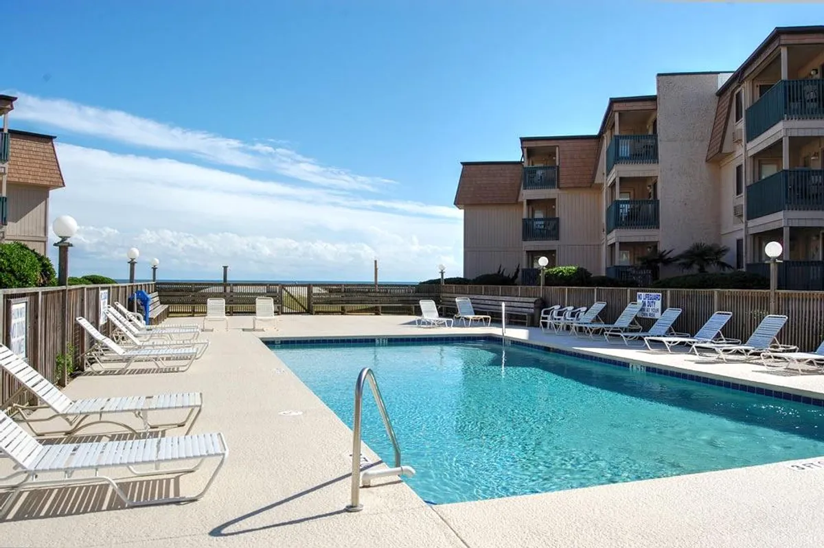 A sparkling resort swimming pool surrounded by lounge chairs, with partial ocean views and the condo buildings in the background. The pool area exudes an inviting and communal vibe, hinting at relaxing swims, playful splashing, and poolside socializing—an essential amenity for families.