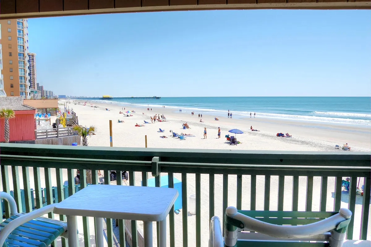A wider exterior perspective taken from a balcony, showing outdoor furniture, people enjoying the beach below, and a distant pier. The setup implies a lively yet relaxing atmosphere, blending personal space with vibrant surroundings.