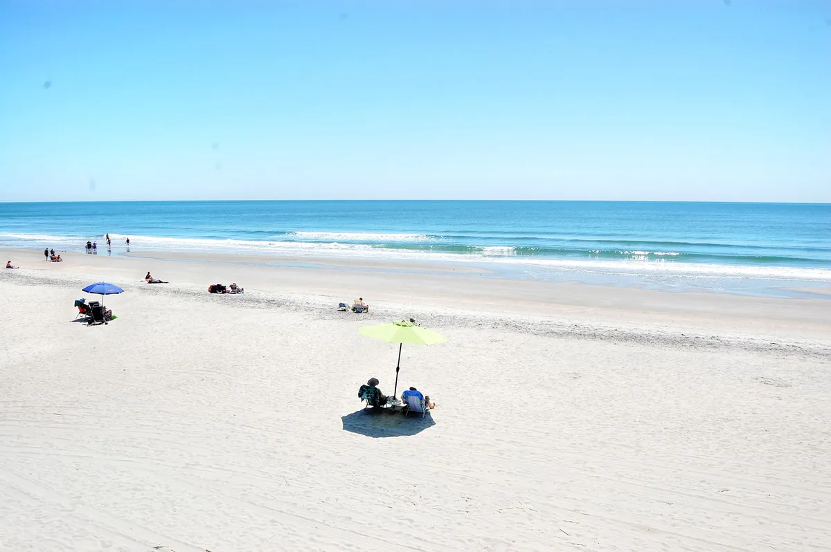 A sun-drenched, wide-angle exterior shot of the beach with gentle waves, beach umbrellas, and couples or individuals walking along the shore. The scene feels tranquil and spacious, inviting visitors to imagine leisurely strolls and shared moments by the ocean.