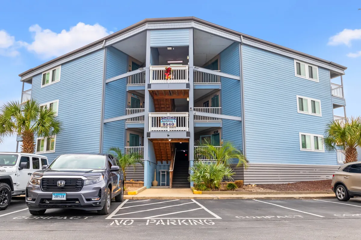 This image showcases the blue building facade of the resort, featuring an inviting entrance with stairs, balconies, and palm trees. The photo captures both the welcoming atmosphere of the property and hints at the social interactions that take place on the balconies and in the parking area.